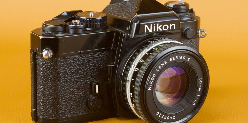 A vintage Nikon film camera with a 50mm f/1.8 lens is displayed against an orange background. The camera features a black body with manual controls and a classic design.