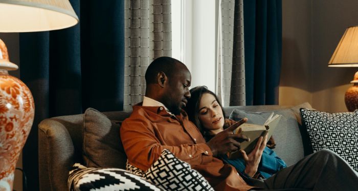 a dark brown-skinned Black man and a fair-skinned white woman reading a book on a couch