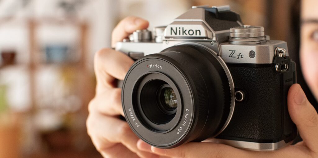 A person holding a Nikon Z fc camera with a 35mm lens. The camera has a vintage design with a silver and black finish. The background is softly blurred, highlighting the focus on the camera.