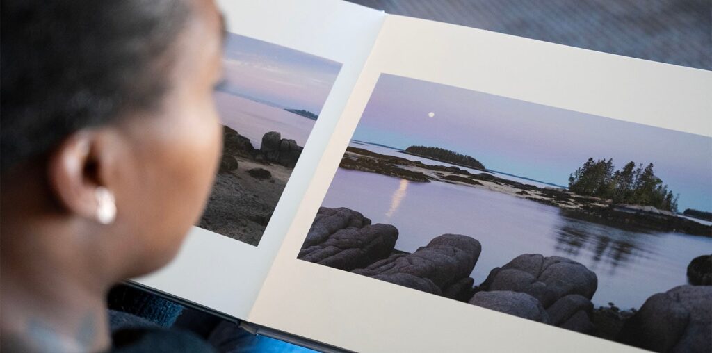 A person is looking at an open photo book displaying serene landscape images of a rocky shoreline and calm waters under a pastel sky. A small island with trees is visible in the distance.