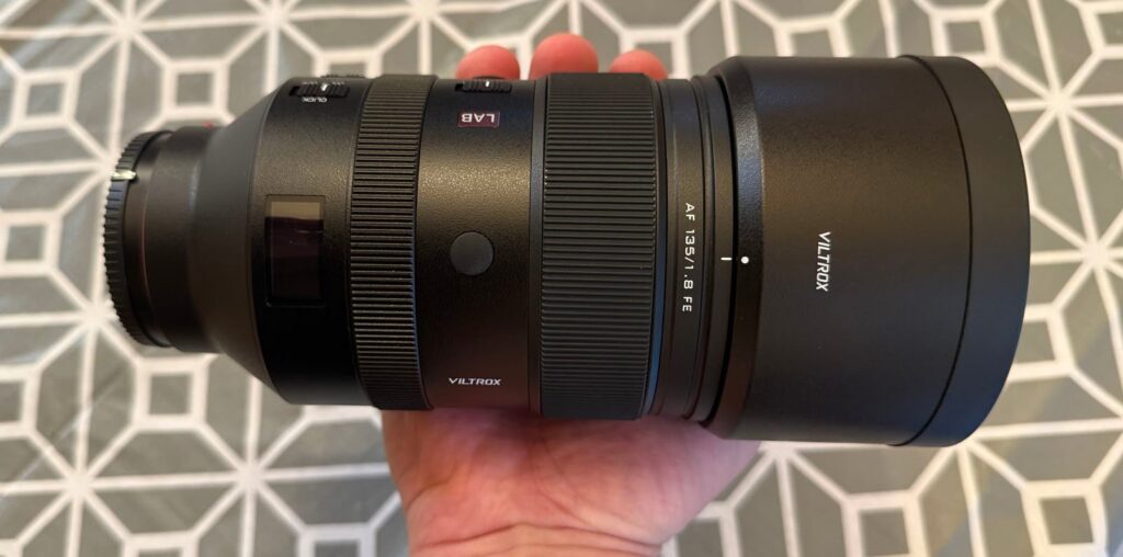 A person holding a Viltrox AF 135mm f/1.8 FE camera lens, with a geometric patterned background. The lens is large and black, featuring various dials and labels.